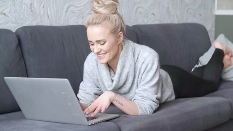 Close up Happy Pretty Young Woman Wearing Casual Clothing. Lying Prone on Sofa with Laptop While Looking at the Camera.