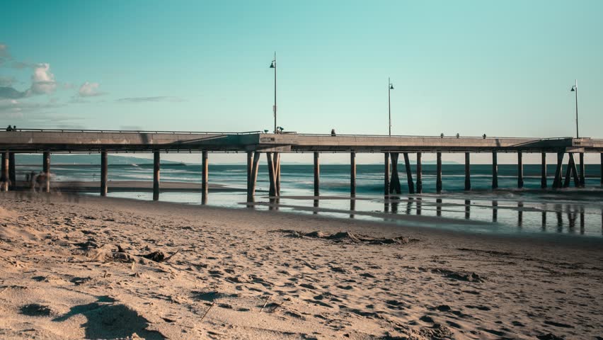 Slow zoom in fast timelapse low angle view of section of Santa Monica pier, Venice beach. Fast motion people passing along sandy beach.