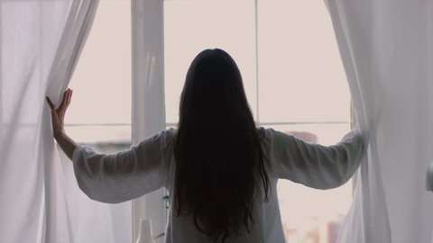 Longhaired brunette girl opening curtains window stretching in morning natural light 4k back view. Sexy woman stands raising arms hands up silhouetted indoors 4k. Freedom femininity fashion clothes
