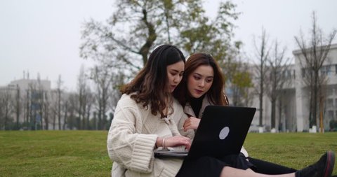 Two Asian female college students in makeup snuggle up in the grass using a computer to study. happy Chinese college students talking and using computer together in the campus 