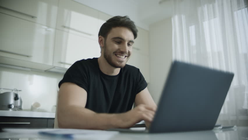 Happy man chatting online on laptop computer at home. Smiling man chatting on laptop at kitchen. Modern people internet lifestyle. Smile male face browsing web on computer | Shutterstock HD Video #1027862591
