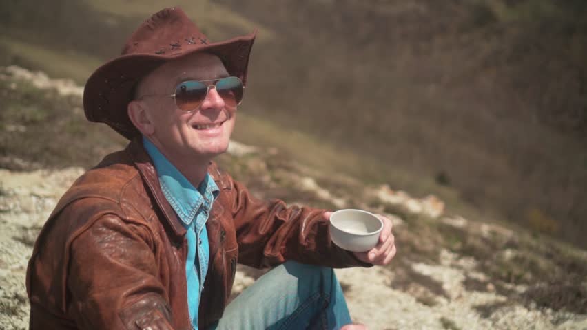 In the mountains sits a man in a cowboy hat, leather jacket, blue jeans and glasses. The man looks at the frame and smiles. Background of mountains and sky.