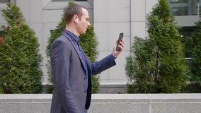 Young businessman walk with wireless earphones and aggressively leads a discussion on a video call on smartphone. A guy in a suit shows his displeasure at a video conference. 4K footage