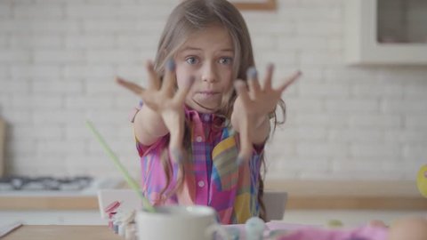 Cute emotional little girl with long hair showing to camera hands in the blue paint, making a grimaces in the kitchen. The child forming heart from her fingers. Concept of creativity