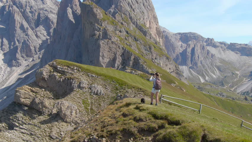 DRONE: Carefree female tourist stops at the scenic mountaintop to take pictures of the beautiful mountain landscape in the Italian Alps. Woman hiking in Dolomites with puppy taking photos of nature.