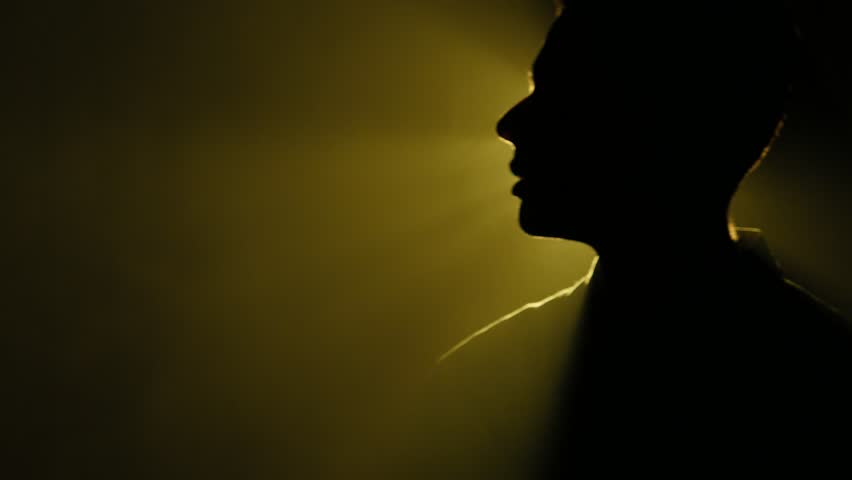 Silhouette of young man walking and looking around in the dark smoky room. Bright yellow lamp behing the man