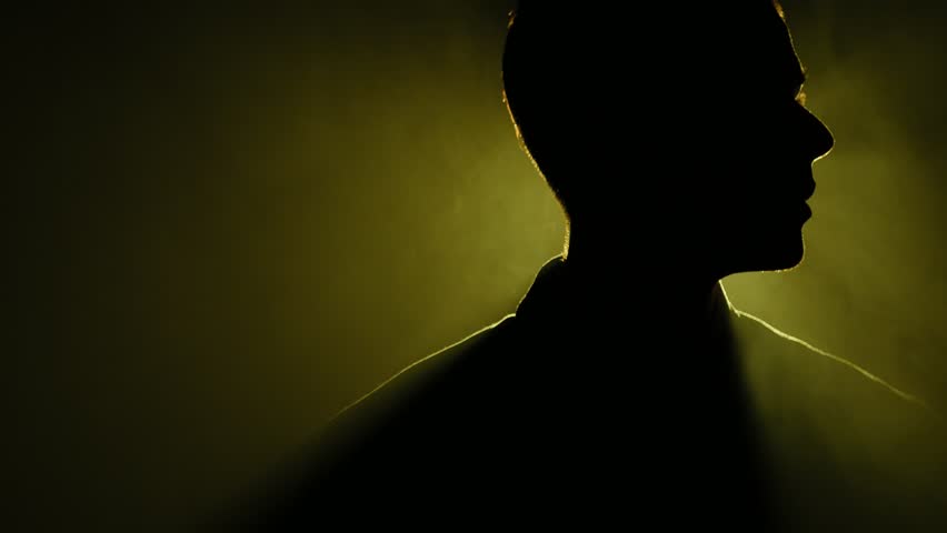 Silhouette of young man standing and looking around in the dark smoky room. Bright yellow lamp behing the man