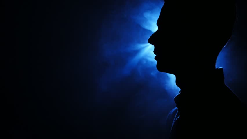 Silhouette of pensive guy standing in the dark smoky room. Bright blue lamp behing the man
