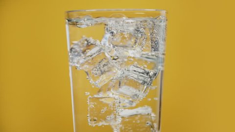 Glass full of cold sparkling water with lot of ice cubes on yellow background, transparent fizzy drink. moving bubbles in soda slow motion. Summer party
