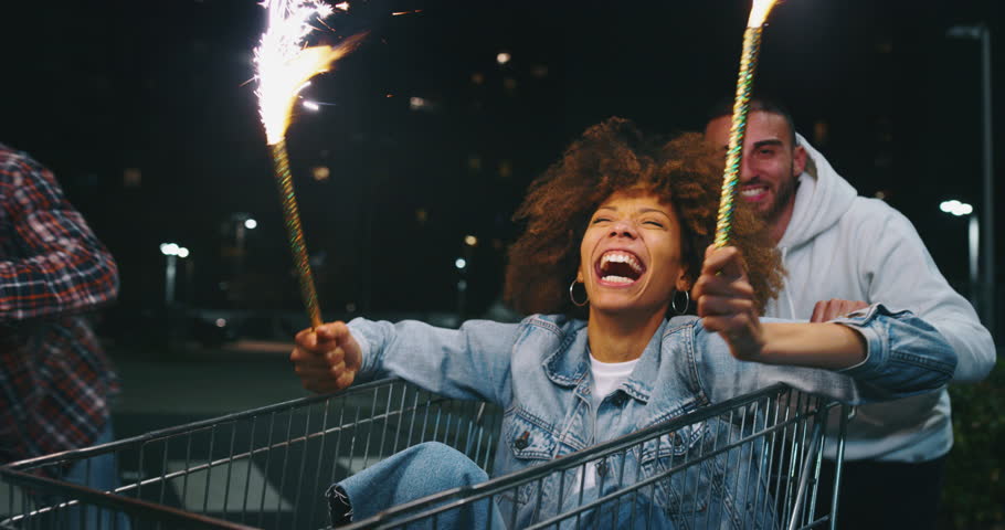 Slow motion close up of group of young friends of different ethnicities are having fun together racing on shopping carts and sparklers at supermarket parking at night.