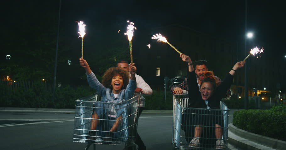 Slow motion of group of young friends of different ethnicities are having fun together racing on shopping carts and sparklers at supermarket parking at night.