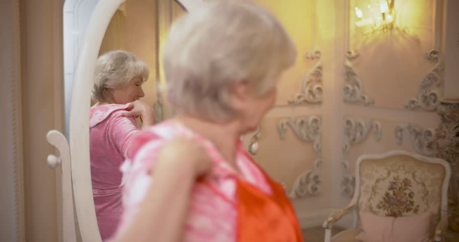 Bright elderly woman in front of mirror with a dress in hands preparing for going out