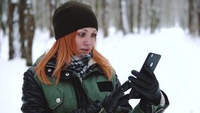 Girl in winter clothes uses a smartphone, a snowy forest. digital technology in cold weather, battery discharge, social networks, watching videos, making bets, laughing and rejoicing, smile on face