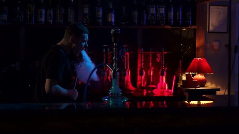Confident man resting in lounge restaurant on against black background, smoking hookah. A young man smokes a hookah in a hookah bar on the background of a bar with hookahs in the dark, slow motion.