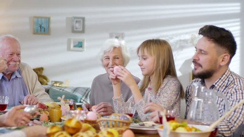 Panning shot of family with children, parents and grandparents talking and smiling while having Easter dinner together, girl giving egg to granddad
