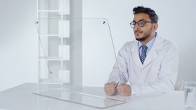 Lockdown of Arab male doctor in lab coat swiping on transparent monitor of futuristic computer while sitting at hi-tech desk in office. Video suitable for adding AR graphics