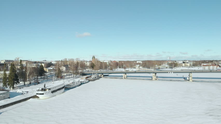 Aerial, tilt up, drone shot, above Pielisjoki, bypassing large boats, towards a bridge, at the coast of Joensuu, towards the cityscape, on a sunny, winter day, in Joensuu, North Karelia, Finland