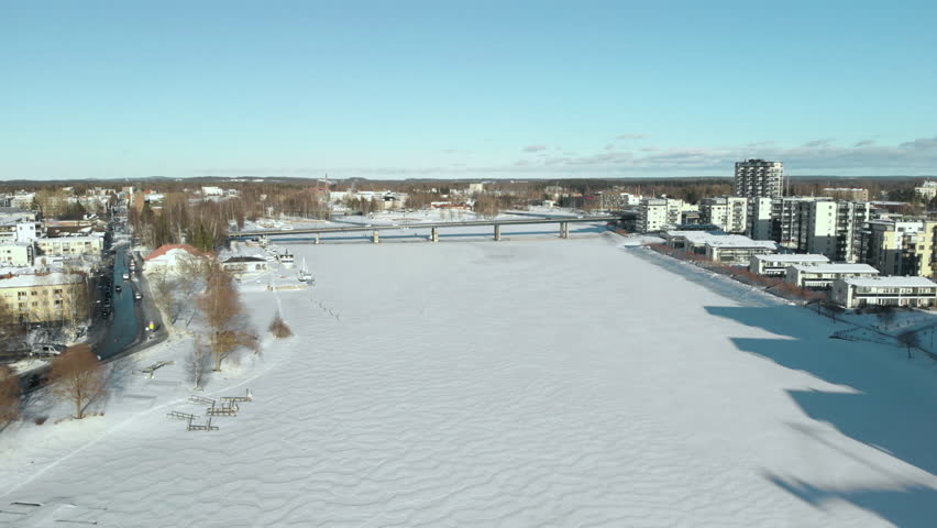 Aerial, drone shot, above Pielisjoki, towards a bridge and apartment buildings, in the cityscape, on the coast of the river, on a sunny, winter day, in Joensuu, North Karelia, Finland