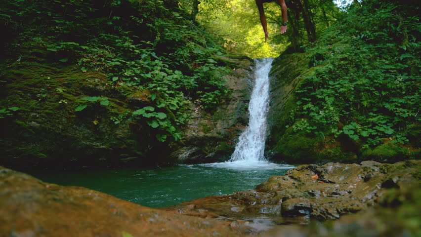 SLOW MOTION: Young Caucasian man jumps into the refreshing stream water on hot summer day. Athletic male tourist jumping into the pond emerald being filled by a forest river. Glassy water splashes