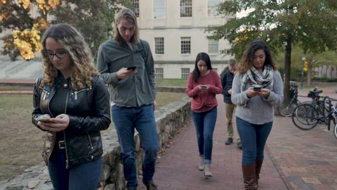 Multi ethnic group of female and male college students walking outdoors on a university campus all on their mobile phones never looking at anything but their smart phones