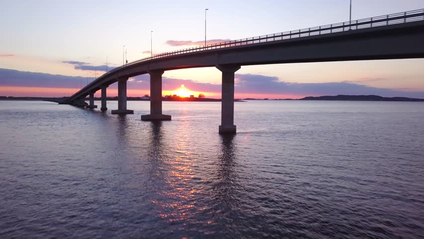 Cinematic drone footage , flying under bridge in a middle of a sunset. pink and red skies, and sun going down in the water