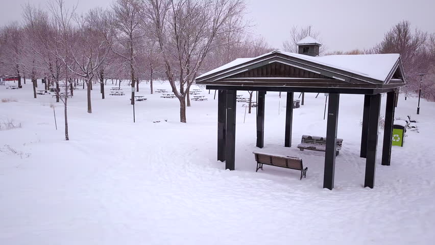 Cinematic drone / aerial footage circling an open hut in a park over a snowy landscape, frozen lake, pathway and dry trees during winter season.