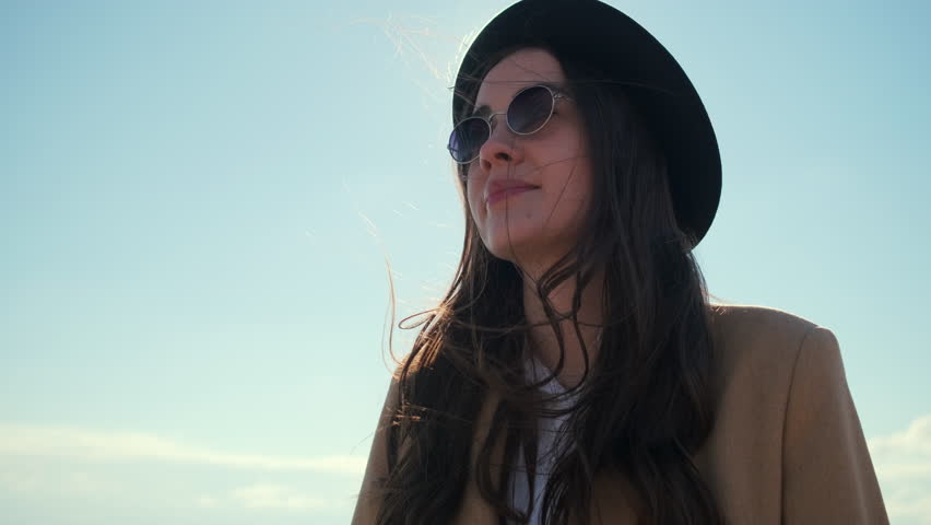 Young charming girl is rejoicing good sunny weather and standing outdoors. Tilt up view in her face with sunglasses in front clear blue sky and sun