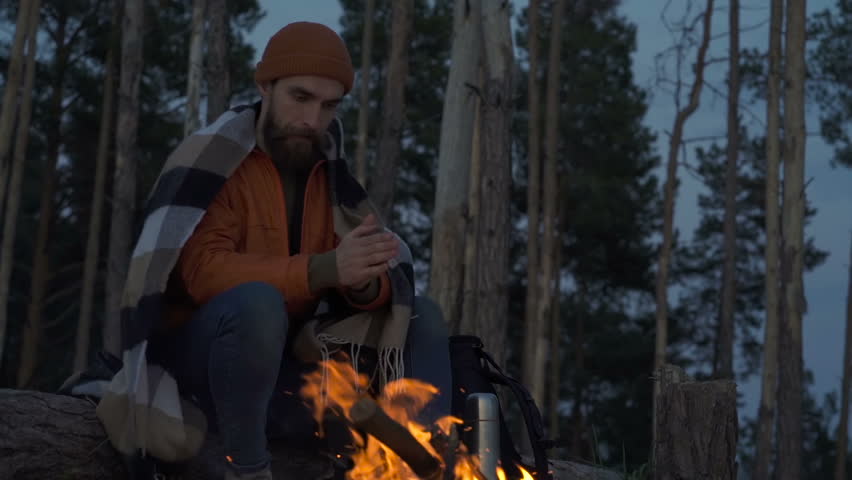 Man with plaid on him sitting near the bonfire and looking at it. Evening time.