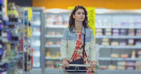 Pretty young woman walks through the supermarket hall with a cart and smiles. Girl chooses goods in the store and carries a trolley.