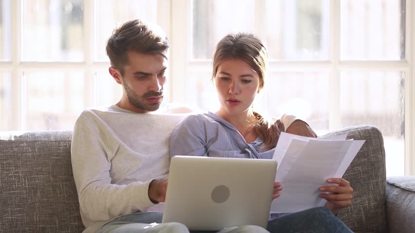 Serious couple talking holding papers discuss pay domestic bills on laptop at home, young family having problem with online banking payments disputing over high taxes expenses worried about loan debt | Shutterstock HD Video #1028009564