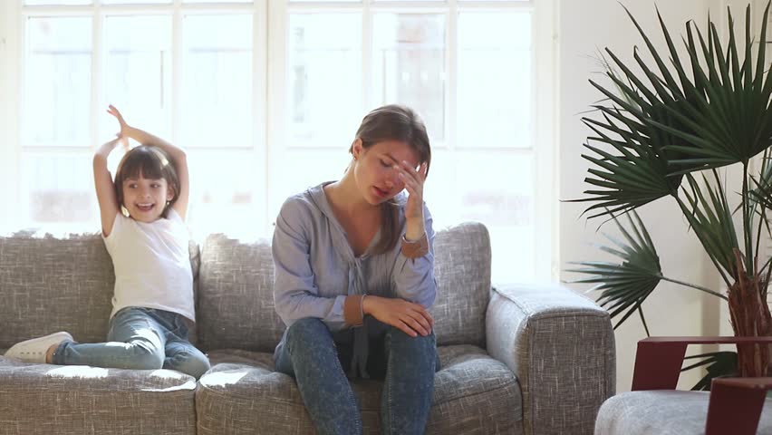 Tired upset single mother feeling stressed about active naughty child daughter jumping on sofa, exhausted sad woman young mom depressed with difficult kid girl frustrated desperate having headache | Shutterstock HD Video #1028009588