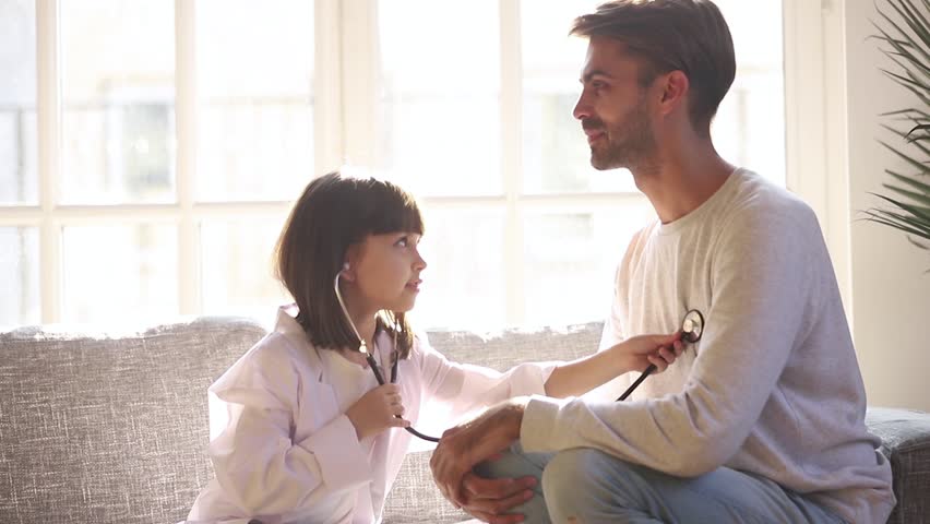 Cute small child daughter dressed in white coat holding stethoscope listening to father patient at home, funny little kid girl playing game as doctor pretending nurse having fun with dad sit on sofa Royalty-Free Stock Footage #1028009591