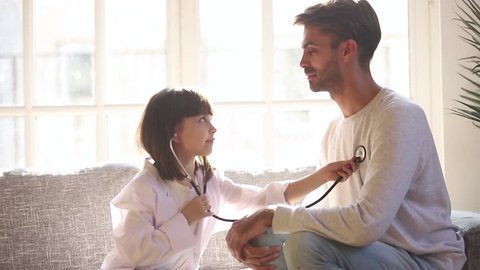 Cute small child daughter dressed in white coat holding stethoscope listening to father patient at home, funny little kid girl playing game as doctor pretending nurse having fun with dad sit on sofa