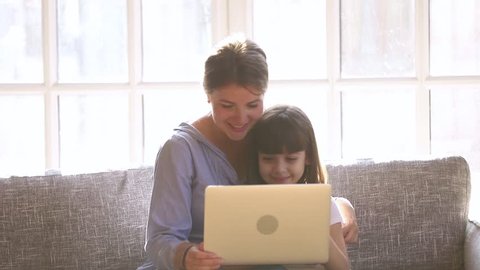 Happy mom with little child daughter having fun enjoying using laptop looking at screen, mother embracing cute small kid girl laughing talking watching cartoons make video call sit on sofa at home