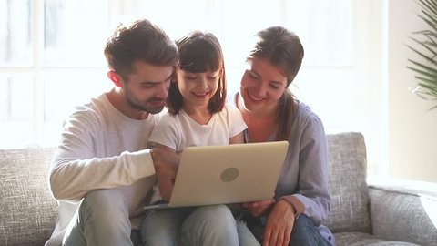 Cute little child daughter laughing looking at laptop screen watching cartoons with parents at home, happy mom dad and small kid girl using computer together enjoying do internet shopping talking 
