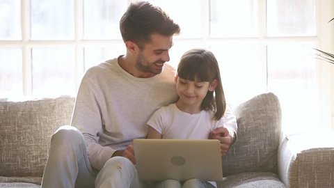 Happy dad and little kid daughter laughing having fun with computer laptop sit on sofa at home, young father embracing child girl talking teaching using modern device together on family leisure