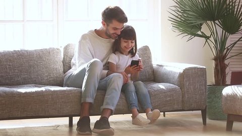 Happy family father and kid daughter using mobile app taking selfie looking at smartphone sit on sofa at home, smiling dad with cute child girl make self portrait snapshot posing for photo on phone
