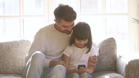 Happy loving father and little daughter using smartphone looking at cell phone screen sit on sofa, cute girl holding cellphone with parental control, smiling dad with child play mobile apps games 
