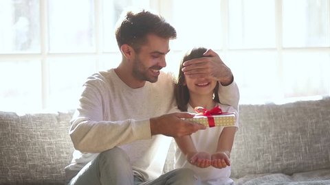 Happy cute daughter receiving gift box from loving father embracing little child girl sit on sofa at home, caring dad congratulating funny little kid making birthday present surprise hugging cuddling