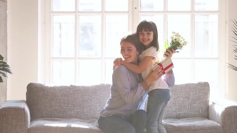 Little cute daughter make surprise for happy mom congratulate mum with mothers day presenting gift box and flowers bouquet, excited mommy receive present from child girl hugging thanking kid at home