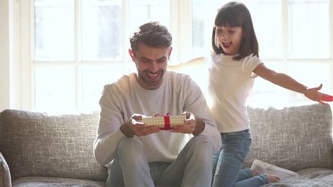 Cute kid daughter make surprise present covering eyes of happy dad receive gift box sit on sofa, little child girl congratulating parent smiling excited daddy on fathers day birthday embrace at home
