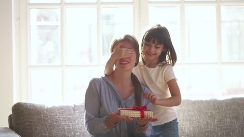 Little cute child daughter closing mom eyes congratulate happy mum with mothers day concept holding gift box, excited mommy receiving present from kid girl make surprise hugging sit on couch at home