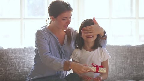 Loving mom make surprise closing eyes of cute child daughter give gift box embrace happy little girl, caring mother congratulate small kid with birthday prepare present on holiday hug sitting on sofa