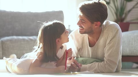 Loving dad and little kid daughter draw with pencils laying on warm floor in living room lit with sunlight, father coloring picture with child girl talk at home enjoy creative activity play together