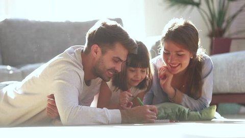Happy family young parents drawing coloring picture with pencils helping cute child daughter enjoying talk play laying on warm floor at home, mom dad and kid girl having fun in living room on leisure