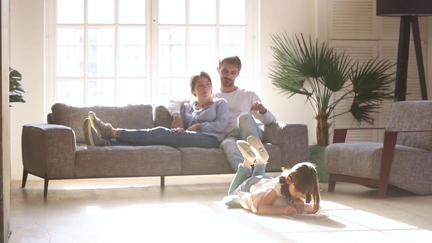 Happy family leisure at home concept, couple parents relaxing talking on sofa couch in comfort living room lit with light while little kid child daughter enjoy activity playing drawing on warm floor Royalty-Free Stock Footage #1028009687