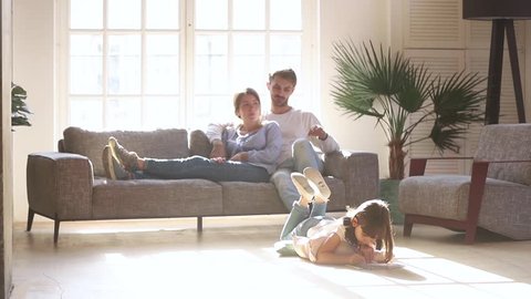 Happy family leisure at home concept, couple parents relaxing talking on sofa couch in comfort living room lit with light while little kid child daughter enjoy activity playing drawing on warm floor