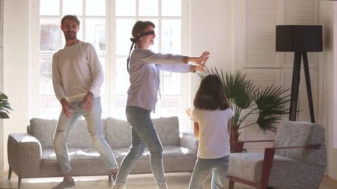 Happy family having fun laughing playing tag touch hide and seek game in living room, blindfolded mom catching child daughter and dad at home, young parents with kid girl enjoy activity together