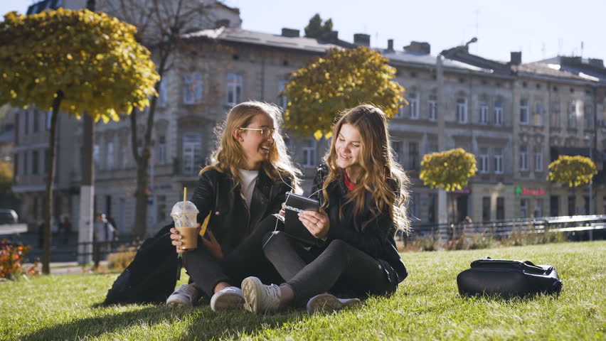 Attractive teen girlfriends using the tablet computer laughing at fun pictures on the tablet sitting outdoors in the park in sunny weather, 4k