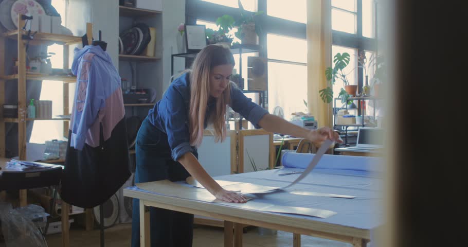 Young woman cutting fabric by edge of paper pattern while making new item for seasonal fashion collection | Shutterstock HD Video #1028015660
